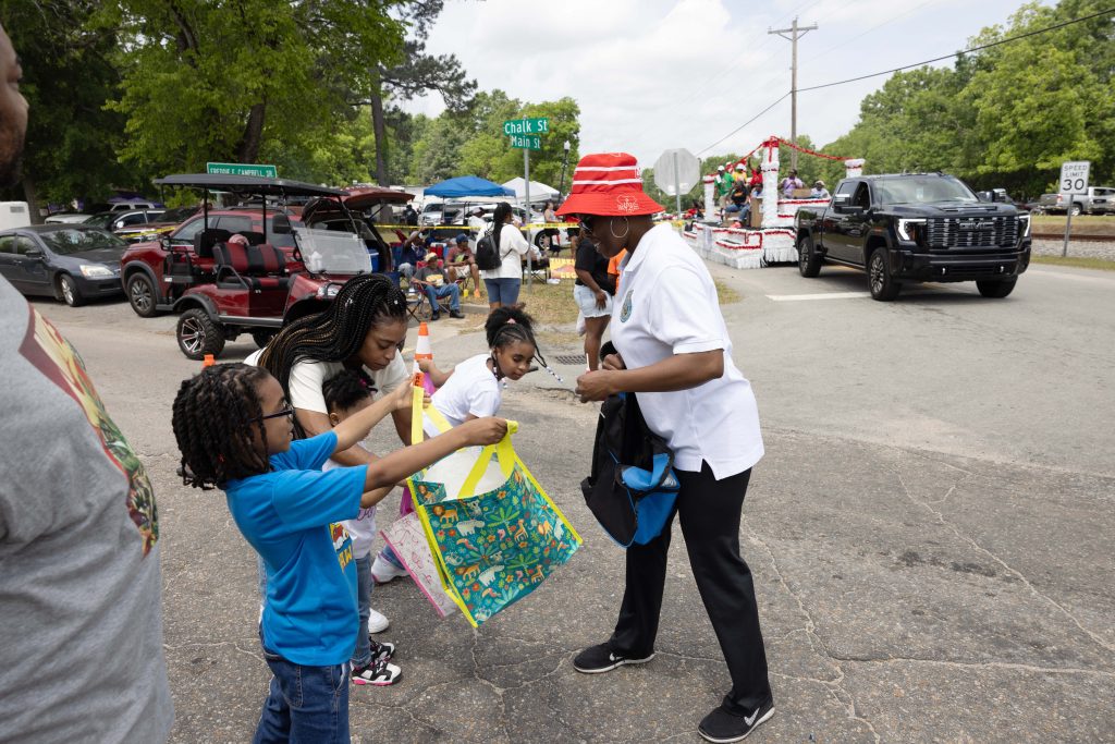 A woman (Tammie Fields) in a red-and-white bucket hat hands out candy to two children holding colorful bags at a parade, with other people, vehicles, and a parade float visible in the background.