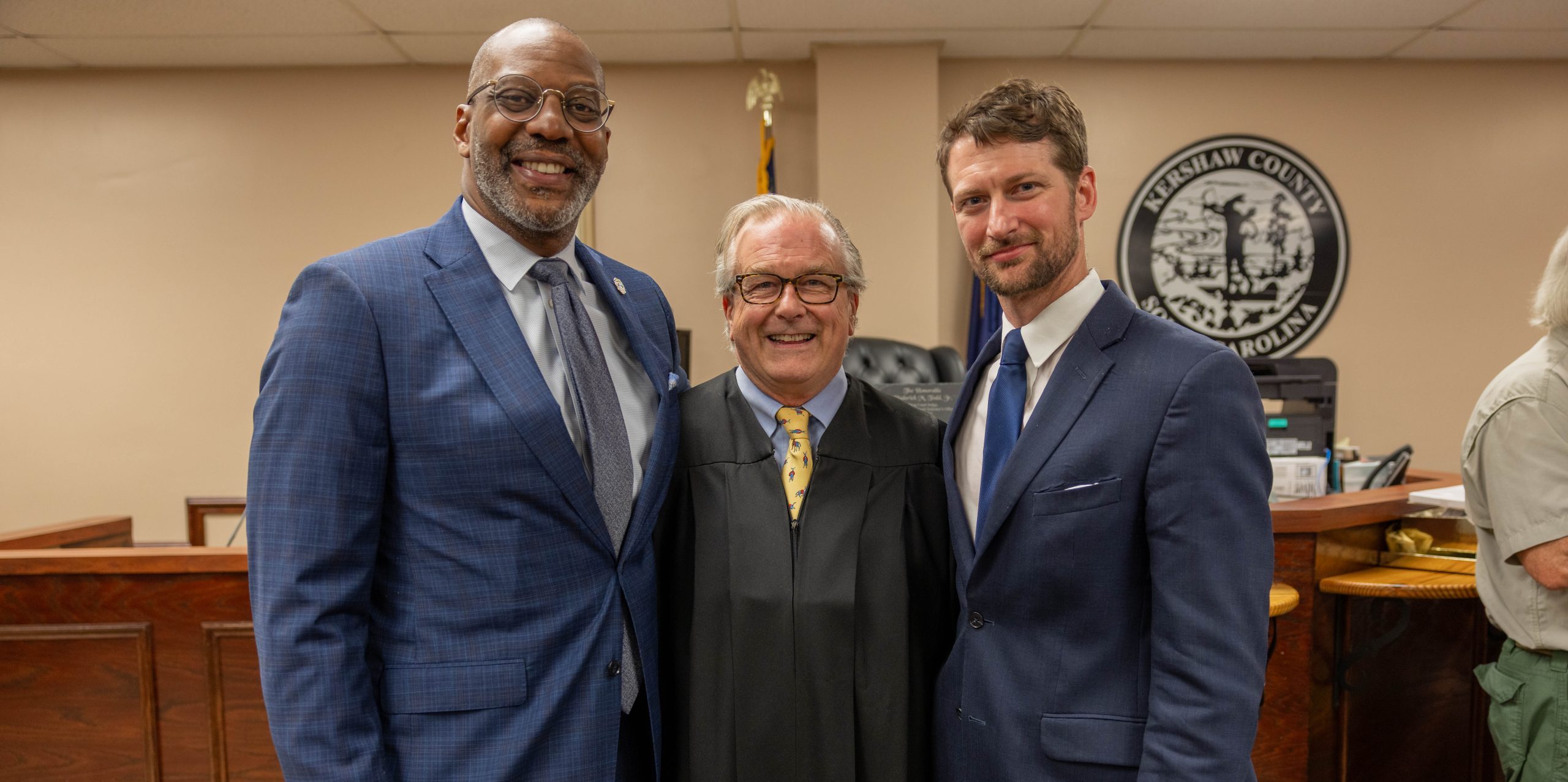 Solicitor Byron Gipson, Judge Roderick Todd, and Judge John Kornegay smile for a photo. Solicitor Gipson and Judge Kornegay are both wearing blue suits, while Judge Todd is dressed in a yellow tie and black judicial robe. In the foreground, a judge's bench is visible, along with a black and white Kershaw County seal on a tan wall.