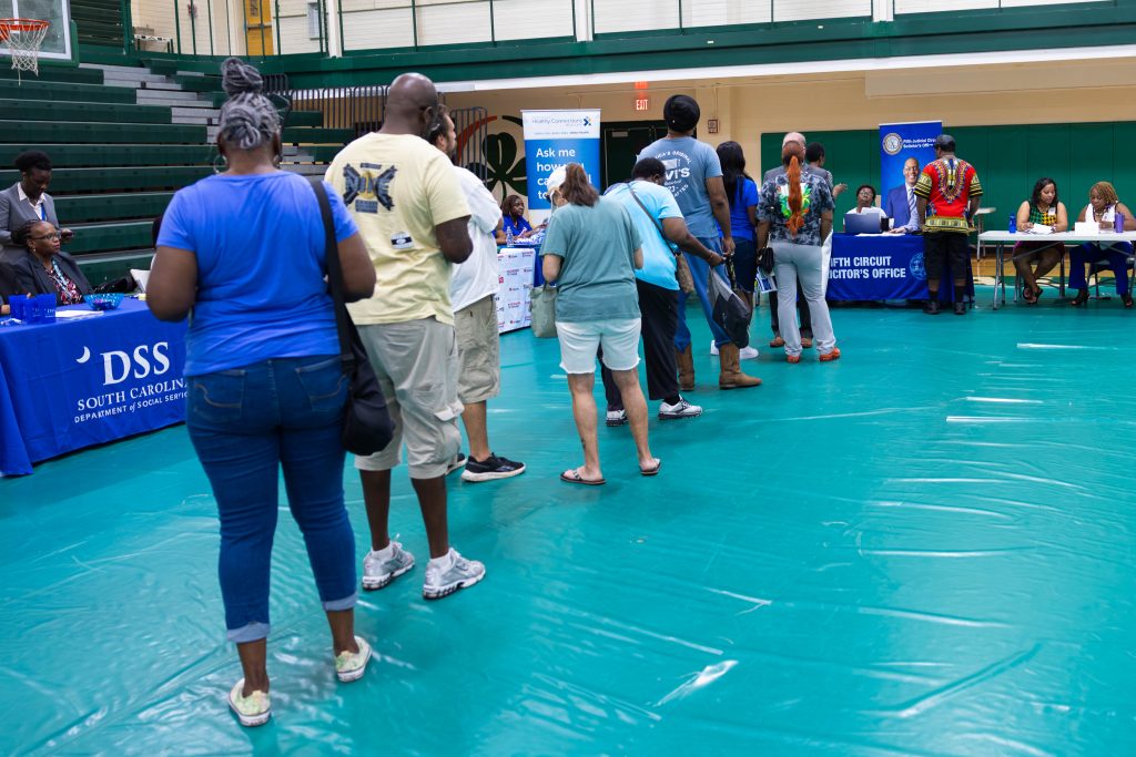 People are standing in line at the Pathways to Possibilities event in the gymnasium of Eau Claire High School. A teal tarp covers the floor, surrounded by forest green bleachers and tan brick walls. In the background, vendor tables are set up.