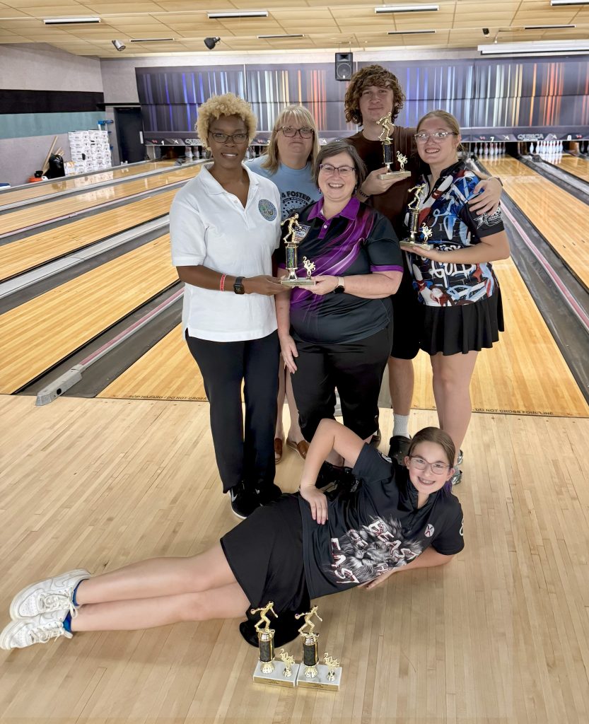 The Fighting Fifth bowling team poses for a group photo. One team member lies on her side with two trophies in front of her, while the other team members stand behind her, holding trophies. In the background, you can see the bowling lanes.