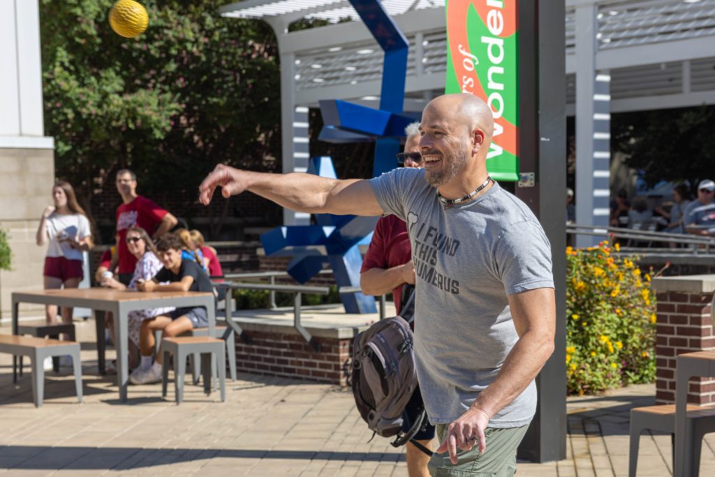 Justin Martin throws a yellow ball while wearing a grey shirt that says, "I Found This Humorous." In the background, a man stands behind him, and several people are seated at a picnic table.
