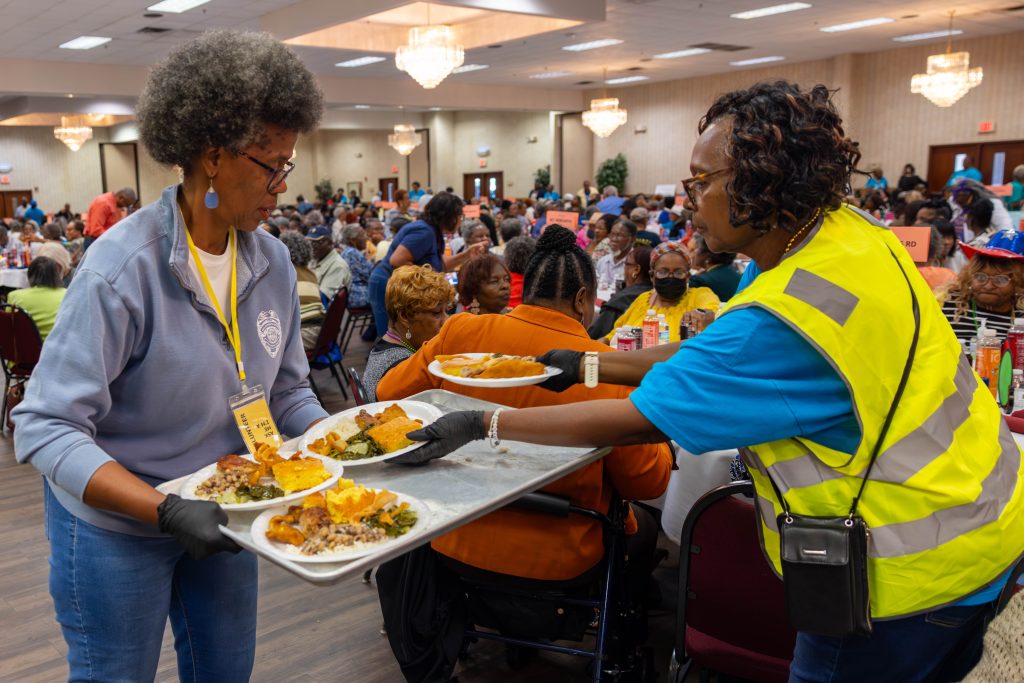 Victim Witness Director Jacquelyn Smith is holding a tray covered with plates of food while a woman in a blue shirt and a yellow reflective vest passes the plates around. Jacquelyn is dressed in a pastel blue quarter-zip sweatshirt and jeans. In the background, people are seated at round tables in a conference room.