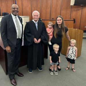 Two men take a photo with a woman and her three children. Her youngest child is on her hip. They are in a courtroom.