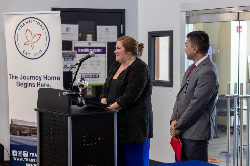 Assistant Public Defender Kieley Sutton speaks into a microphone at a black podium. Next to Kieley is Ph.D. Candidate Dylan Wong. They are both smiling. Kieley is wearing a black top, a cardigan, and royal blue pants. Dylan is wearing a grey suit, a white button-up shirt, and a red tie. He is holding a red folder as he looks at Kieley speak. Behind them is a Transitions Homeless Center pull-up banner, featuring its logo, the phrase "The Journey Home Begins Here," an image of the building, social media icons, and contact information. Next to the banner is a black door covered in white posters, along with a window and glass doors that look into a room.