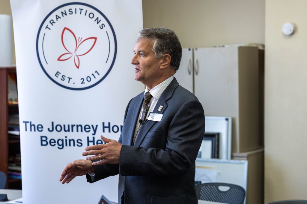Craig Currey, CEO of Transitions Homeless Center, speaks expressively using his hands. He is dressed in a grey suit and a patterned tie, with an army pin on his lapel and a name tag beneath it. Behind him is a pull-up banner from the Transitions Homeless Center featuring its logo at the top, accompanied by the phrase, "The Journey Home Begins Here." Craig is in a pastel yellow room, which has chairs, cabinets, and unidentified objects in the background.