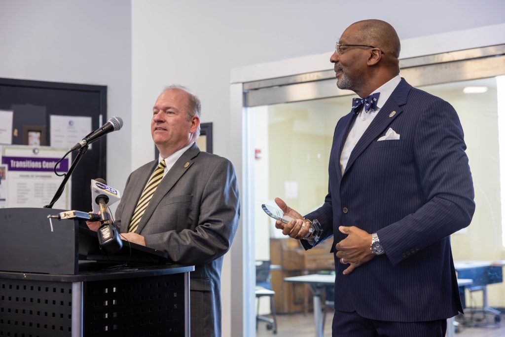 Hans Pauling, director of Diversion Programs and Affiliate Services at the Fifth Judicial Circuit Solicitor's Office, stands at a black podium and speaks into two microphones. He is dressed in a grey suit with a white button-up shirt, a yellow and black striped tie, and he has the pin of the Fifth Judicial Circuit Solicitor's Office on his lapel. Next to him is Solicitor Byron Gipson, who is wearing a blue striped suit with a white button-up shirt, a white handkerchief, and a blue bow tie, along with an office lapel pin. Solicitor Gipson also wears glasses. Behind them are closed glass doors leading to another room, and to their left is a black door adorned with posters.