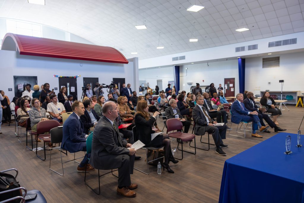 Event attendees are seated in chairs of various colors in Transitions Homeless Center's Day Room. The room features wooden floors and white walls, creating an open atmosphere. Behind the attendees is an enclosed office area for caseworkers, topped with a rounded red roof. The attendees are attentively listening to the speaker, who is not shown in the picture. In the right corner is a table with a blue tablecloth and glass plaques on it.