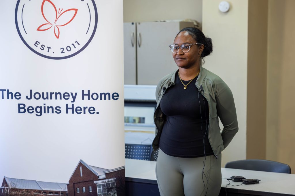 Homeless Court graduate Yasmeen Lewis speaks to the media in a room with pastel yellow walls. Next to her is a white pull-up banner featuring the Transitions Homeless Center's logo and the phrase, "The Journey Home Begins Here." Yasmeen has her hair pulled into a bun and is wearing glasses, a black T-shirt, and a matching olive green workout suit. She also has a gold necklace around her neck. Behind her, there is a table, chairs, and a cabinet.
