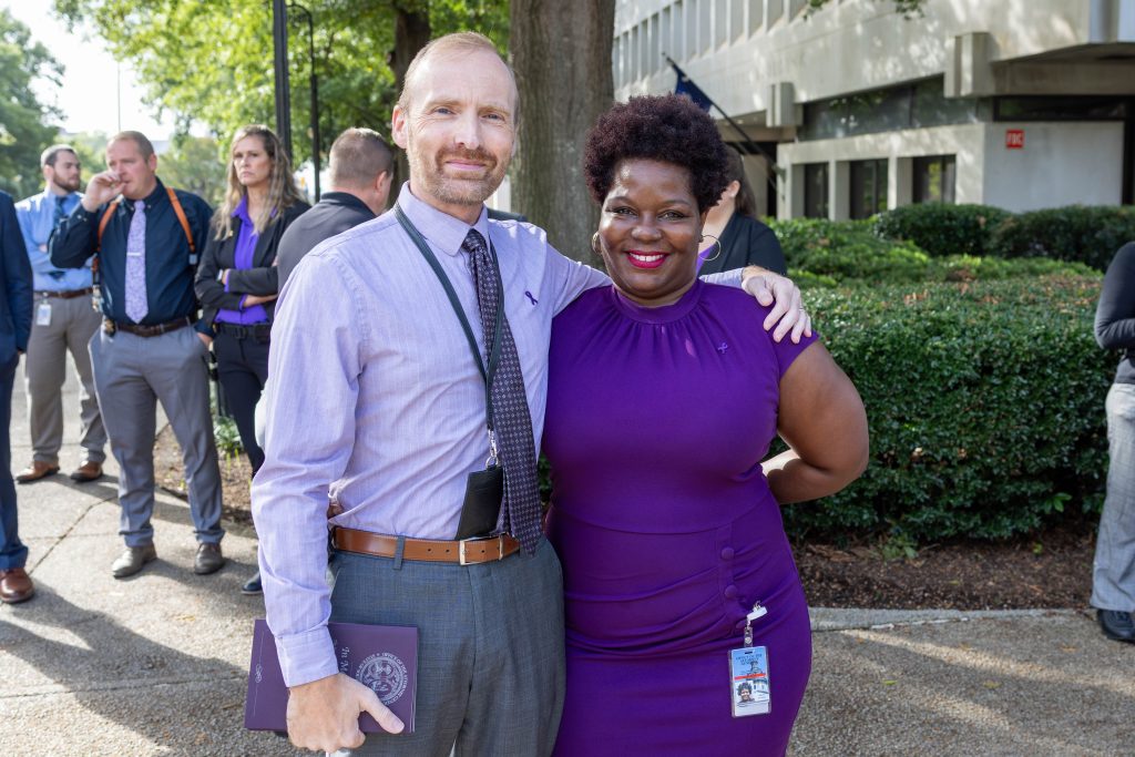 John Dunn, wearing a violet button-up shirt, a patterned tie, an ID lanyard, a brown belt with a gold buckle, and grey slacks, holds a purple booklet in one hand while wrapping his other arm around Nataki Brown. Nataki is dressed in a purple dress with an ID badge clipped to her hip. She has gold hoop earrings and red lipstick. In the background are shrubs, trees, and people.