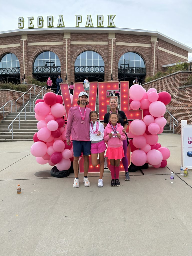 Circuit Deputy Solicitor Dan Goldberg stands with his wife and their two daughters in front of colorful pink balloons and a marquee sign at a baseball stadium. The family is dressed in athletic wear.