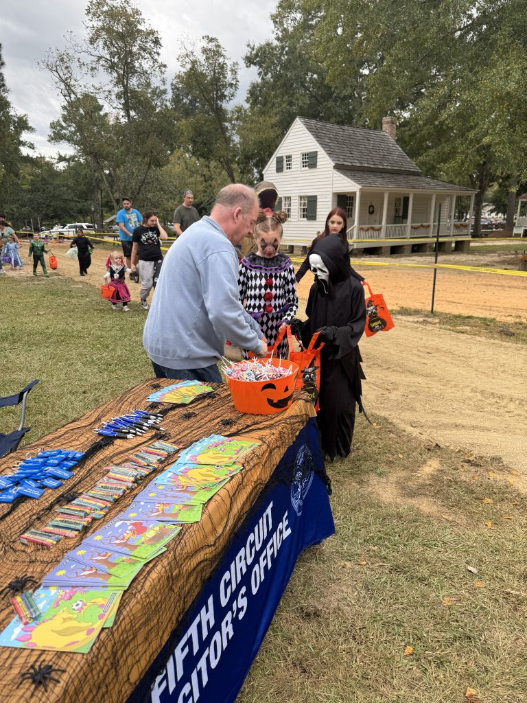 A table draped with a blue tablecloth labeled “Fifth Circuit Solicitor’s Office” is decorated with a brown covering, black spider webs, and plastic spiders. Coloring books and crayons are spread across the table, and an orange jack-o’-lantern candy bowl sits at the end. Hans Pauling leans forward to hand out candy to children dressed as Scream and Pennywise the Clown. A historic white house and trees are visible in the background.