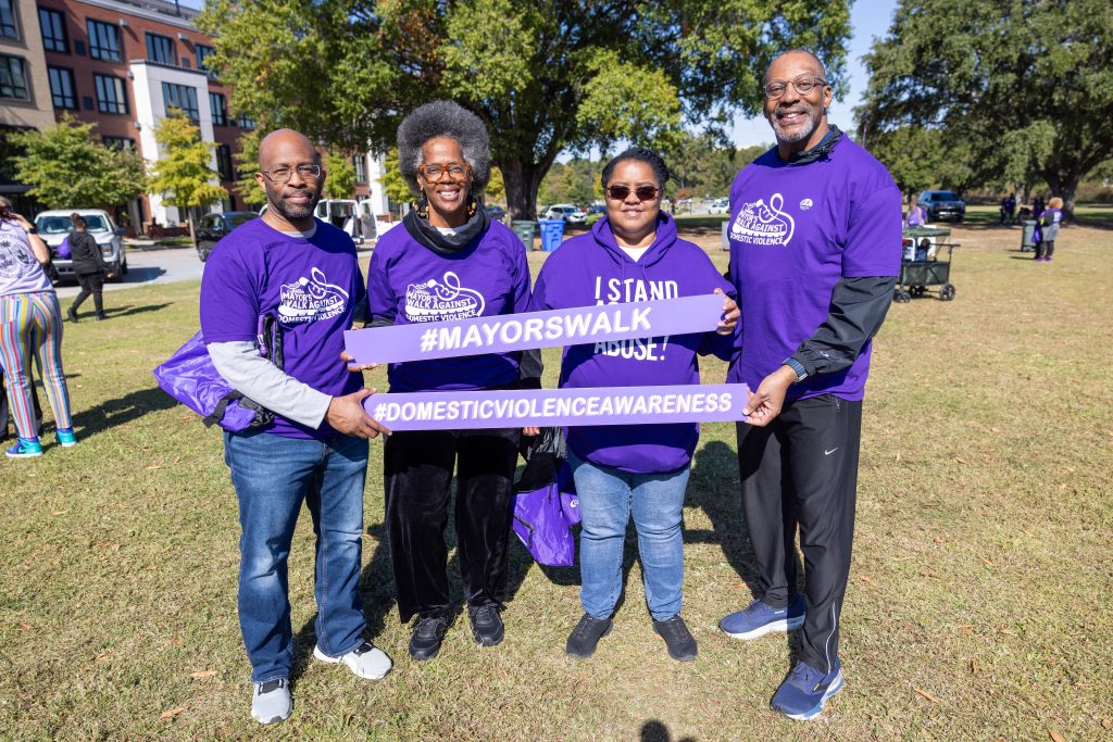 Curtis Pauling, Jacquelyn Smith, Donna Green, and Solicitor Byron Gipson are wearing purple shirts and holding two signs. Jacquelyn and Donna have the top sign that reads, "#MayorsWalk." Curtis and Solicitor Gipson hold the bottom sign that reads, "#DomesticViolenceAwareness."