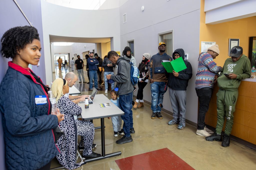 A woman in a blue quilted jacket and red polo stands against a purple wall. Next to her, two people sit at a table checking in attendees. A line of attendees can be seen down the hallway.
