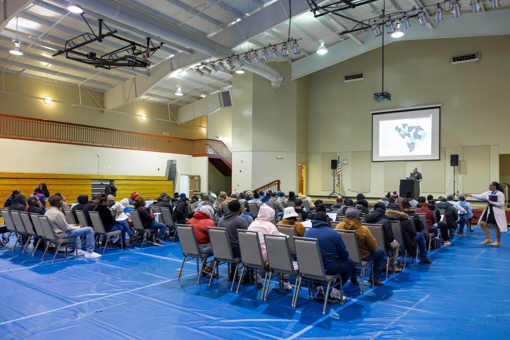 A group of people is sitting in chairs in a gymnasium, listening to a speaker at a podium on stage. Behind the speaker, there is a projection screen. A blue tarp covers the gym floor.