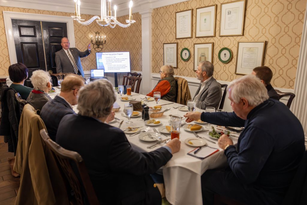Hans Pauling, the director of diversion programs, stands at the front of a small room, pointing to a TV screen that displays a PowerPoint presentation on diversion programs. Attendees are seated around a rectangular table covered with a white tablecloth and plates of food. The attendees are listening attentively while enjoying their lunch. The room has yellow patterned wallpaper, decorative frames on the walls, and a chandelier hanging above the table.