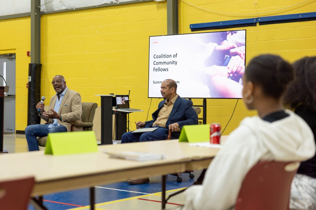 Solicitor Byron Gipson and Judge Milton Kimpson sit in front of high school students, discussing their careers. Behind them, a TV screen displays "Coalition of Community Fellows." The students sit at tables and listen attentively to the guest speakers.